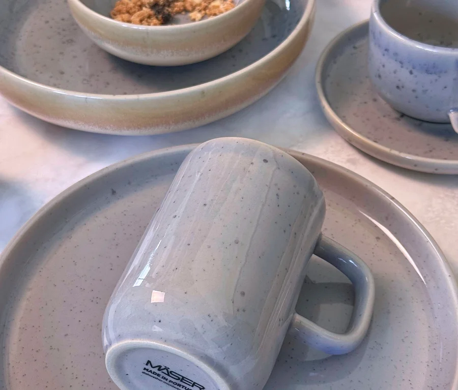 Nahaufnahme eines Mäser Tafelservice »Candy« mit gesprenkelten hellgrauen und beigen Teilen. Ein großer Teller, eine Schale mit Müsli, eine Kaffeetasse mit Untertasse und ein Becher auf einem Teller sind sichtbar.