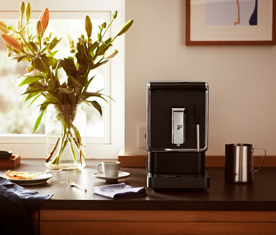 Auf einer Küchentheke steht ein »Esperto Latte« Tchibo Kaffeevollautomat, Anthrazit neben einer Tasse und einer Blumenvase.
