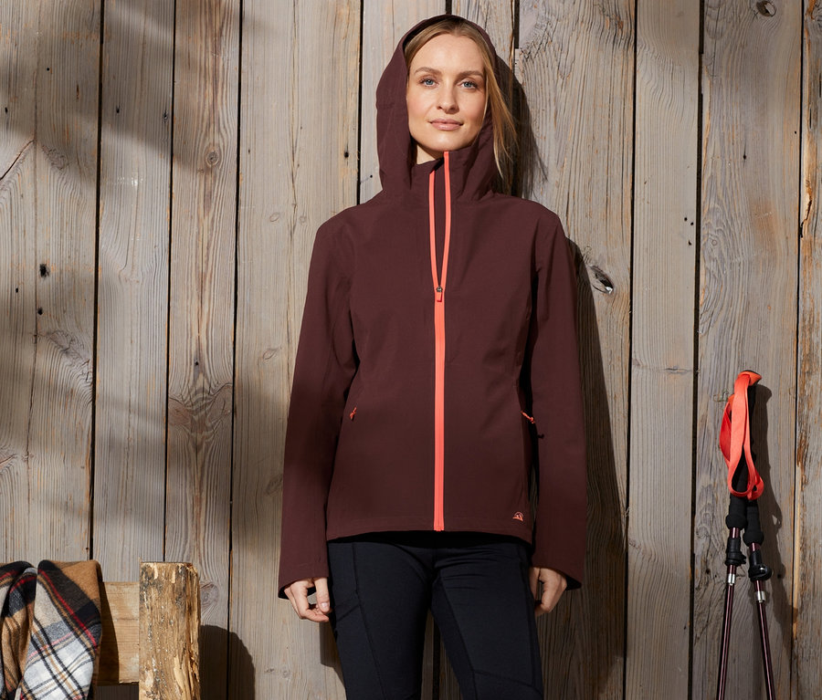 Eine Frau steht an einer Holzwand und trägt eine Funktionswanderjacke mit Kapuze und orangefarbenem Reissverschluss.