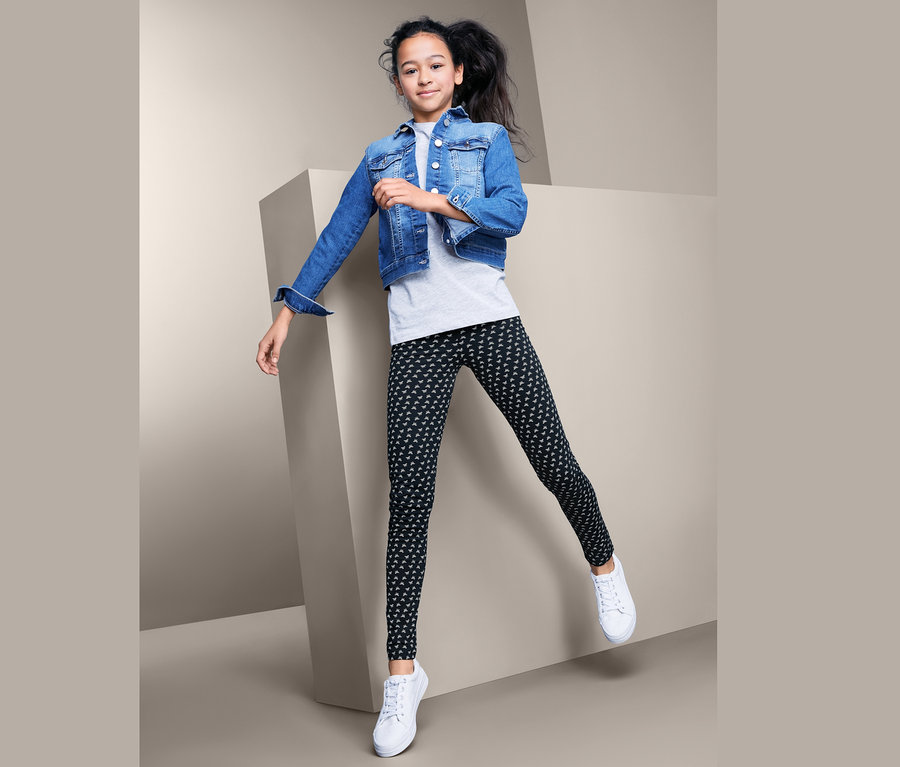 Mädchen springt vor geometrischem Hintergrund, trägt Jeansjacke und 2 Leggings.