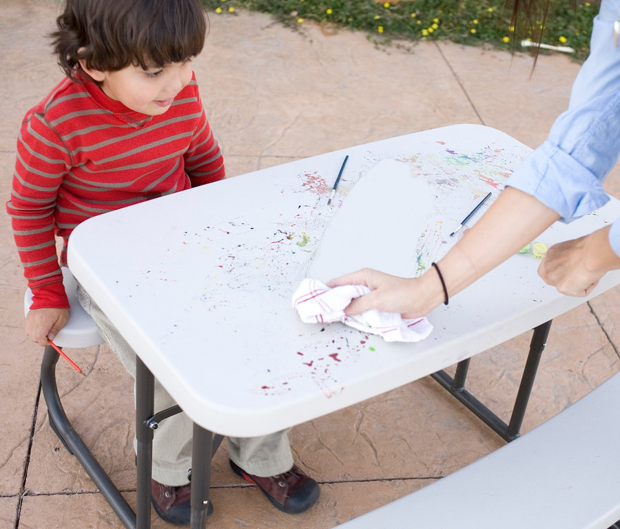 Ein Kind sitzt an einem hellgrauen LIFETIME Kinder-Picknicktisch »80094G« und schaut zu, wie eine erwachsene Person Farbflecken von der Oberfläche wischt.