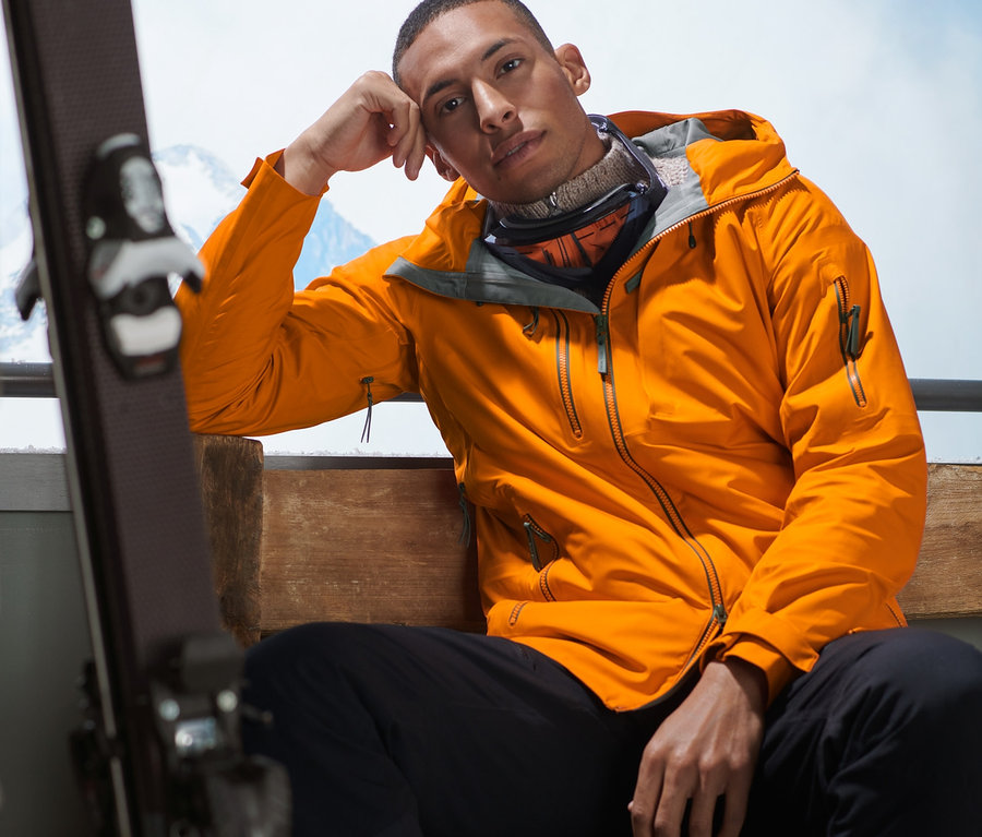 Mann sitzt mit einer orangefarbenen Hardshell-Skijacke auf einer Bank.