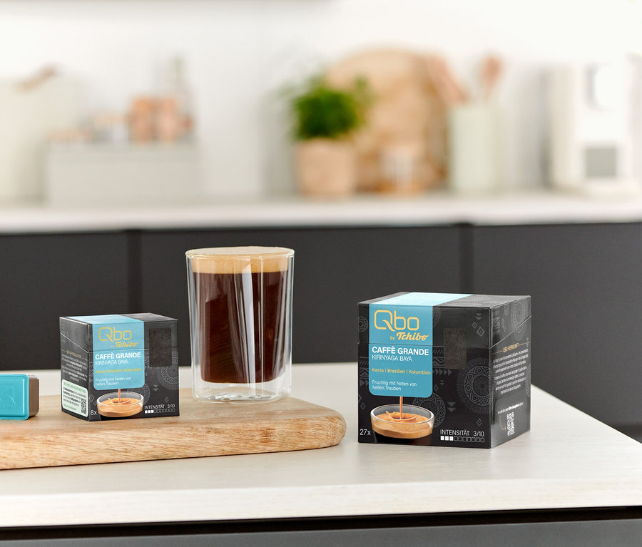 Auf einem Tisch stehen eine Tasse Kaffee, ein Qbo Caffè Grande KIRINYAGA BAYA - 8 Kapseln und ein Qbo Caffè Grande KIRINYAGA BAYA - 27 Kapseln.
