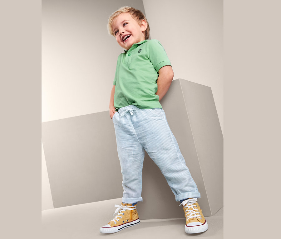 Ein Junge lacht und posiert in einer Baumwoll-Schlupfhose mit Leinen, einem grünen Poloshirt und gelben Turnschuhen.