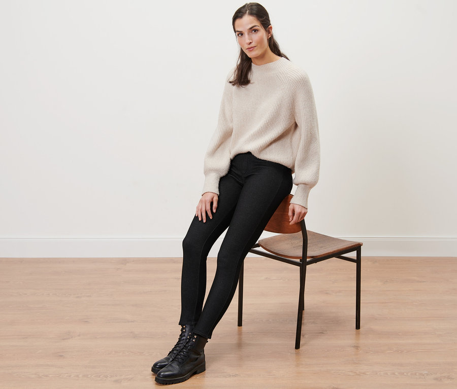 Frau sitzt auf einem Stuhl und trägt gefütterte Leggings in Jeansoptik, einen Pullover und Stiefel.