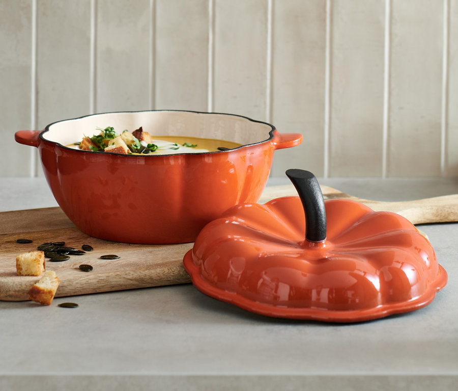 Ein offener Gusseisen-Topf in Kürbisform, gefüllt mit Suppe und Croutons, steht auf einem Holzbrett neben dem Deckel.