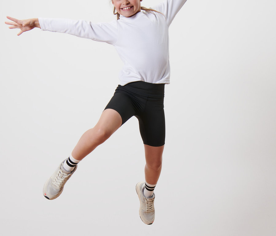 Ein Mädchen springt auf einem Holzsockel und trägt schwarze Sport-Radler und ein weißes Langarmshirt. Sie trägt 2 Kinder-Sport-Radler.