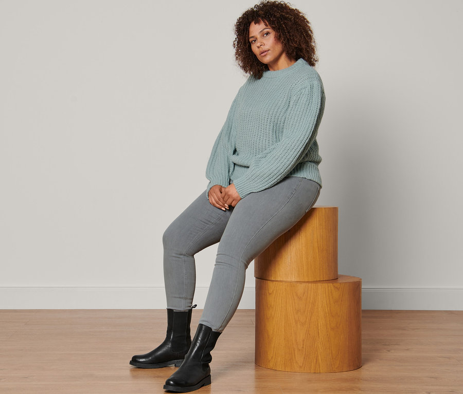 Eine Frau sitzt auf Holzsockeln und trägt einen rauchblauen Grobstrick-Pullover mit Wolle, hellgraue Treggings und Lederboots.