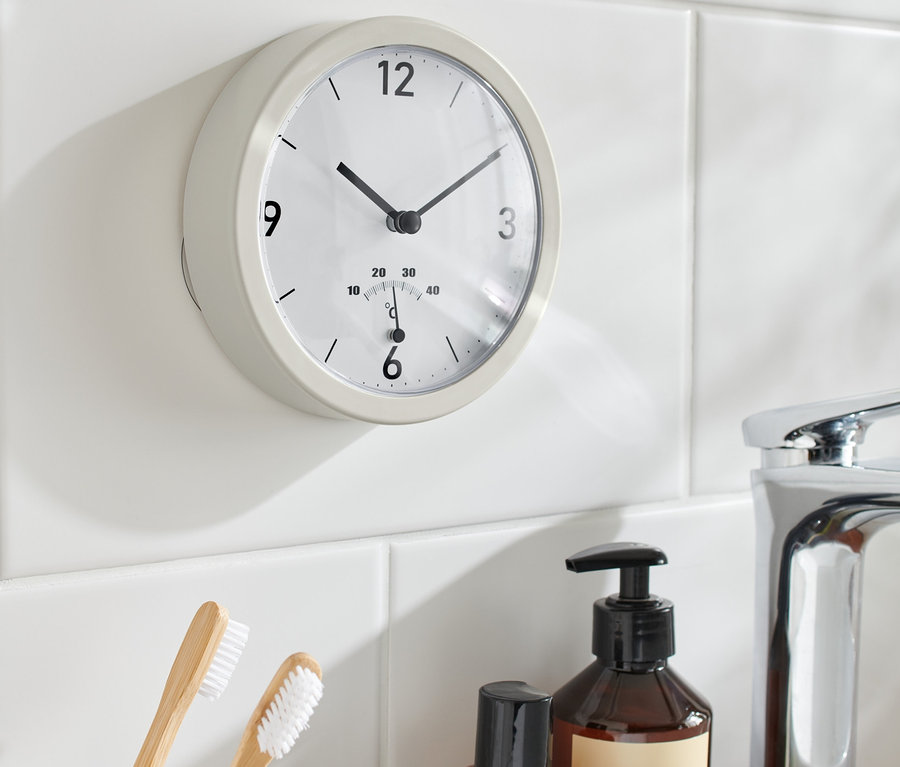 Eine Badwanduhr mit Thermometer hängt an weißen Fliesen neben einem Waschbecken, Zahnbürsten und Flaschen.