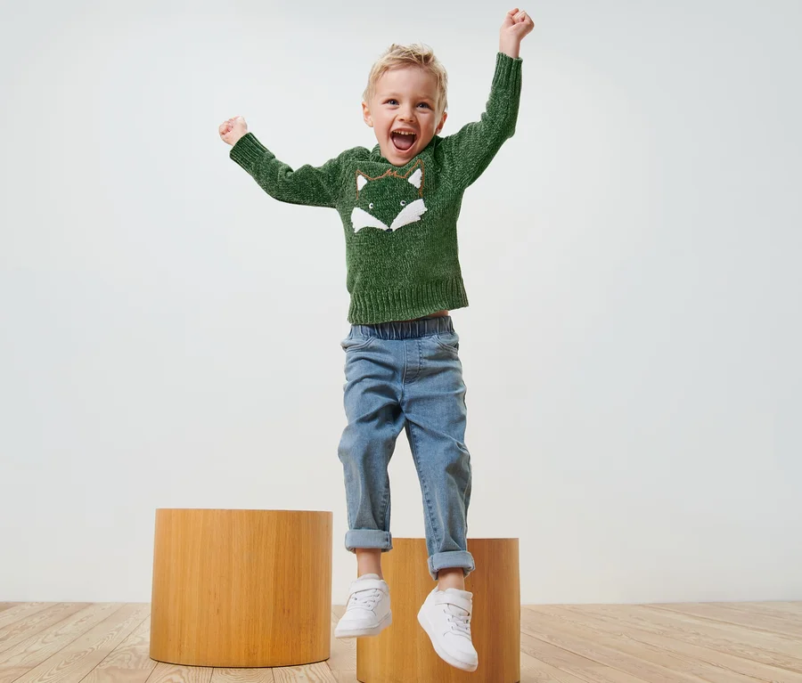 Ein Junge springt auf Holzhockern und trägt einen grünen Chenille-Strickpullover.