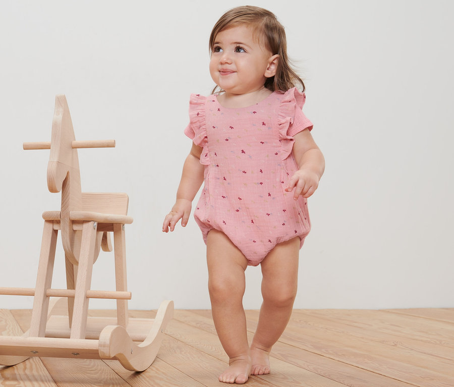 Ein Baby in einem Baby-Strampler steht neben einem Schaukelpferd aus Holz.