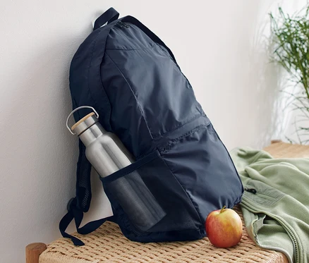 Ein marineblauer, faltbarer Rucksack lehnt an einer weißen Wand auf einer Korbsitzbank. Eine silberne Wasserflasche steckt in der Seitentasche des Rucksacks, daneben liegt ein Apfel.