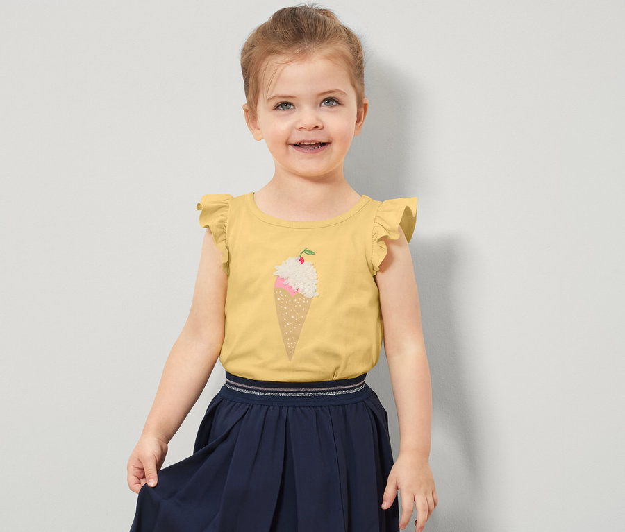 Ein lächelndes kleines Mädchen trägt ein gelbes Kinder-Shirt mit Volantärmeln und einem Eisaufdruck.