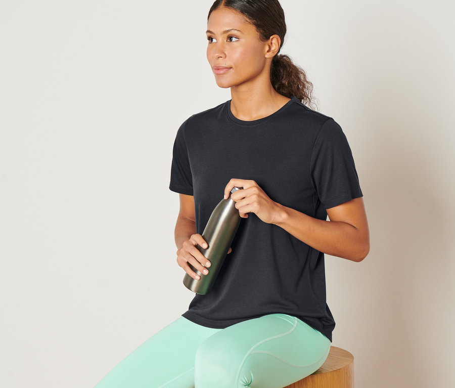 Frau sitzt auf einem Holzhocker und hält eine Metallflasche. Sie trägt ein anthrazitfarbenes Sportshirt.