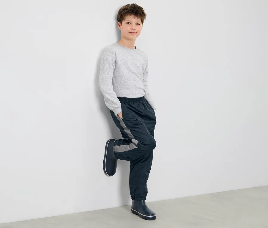 Ein Junge lehnt an einer Wand und trägt eine Kinder-Regenhose und Gummistiefel.