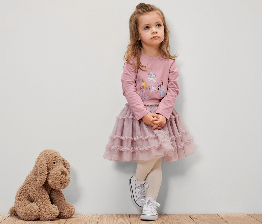 Ein Mädchen in einem rosa Langarmshirt mit Prinzessinnenmotiv, einem rosa Rock und weißen Turnschuhen steht an einer Wand. Neben ihr sitzt ein Plüschhund.