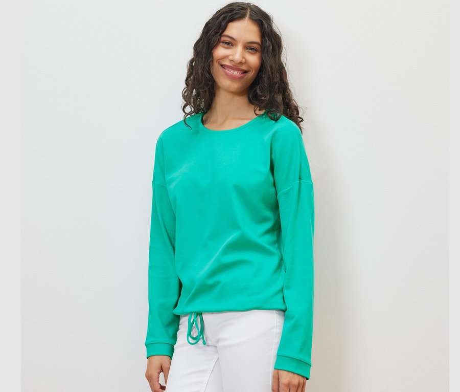 Frau trägt ein Sweatshirt in Aqua und eine weiße Hose.