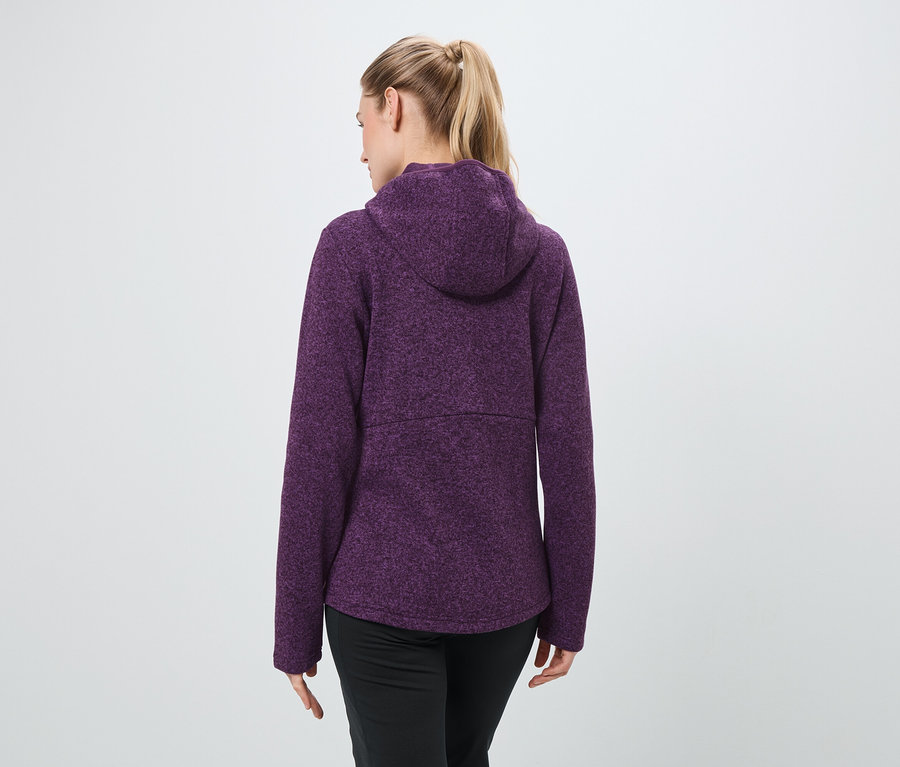 Von hinten ist eine Frau mit blonden, zu einem Pferdeschwanz gebundenen Haaren zu sehen, die einen violetten Kapuzenpullover und eine schwarze Hose trägt.