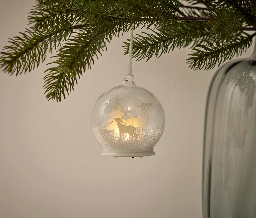 Eine hängende Glaskugel mit weißen Figuren eines Rentiers, eines Baumes und eines Hundes im Inneren, umgeben von weißen Kugeln und beleuchtet, hängt an einem Ast.