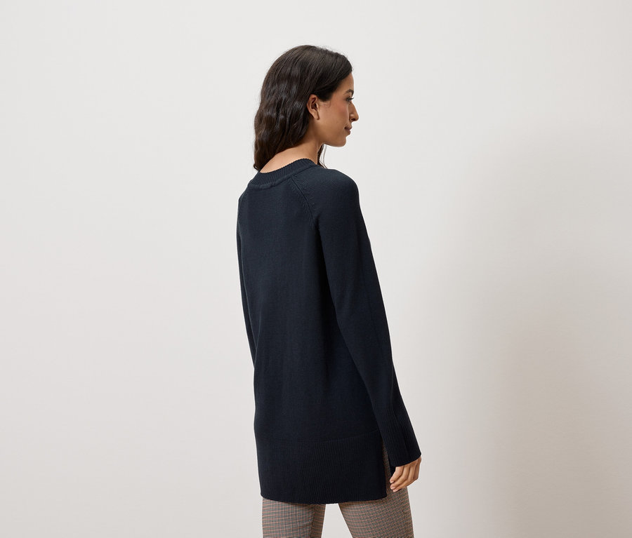 Frau steht seitlich und trägt einen dunkelblauen Pullover und eine karierte Hose.