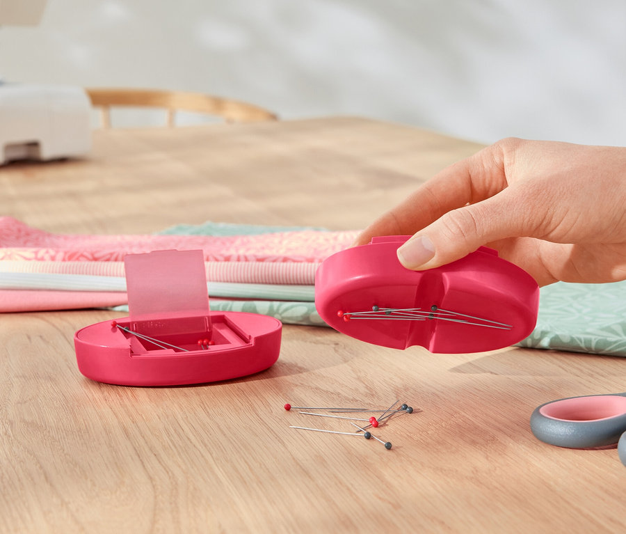 Eine Hand hält ein magnetisches Nadelkissen über einem geöffneten magnetischen Nadelkissen, daneben liegen eine Schere und ein Stoffstapel.