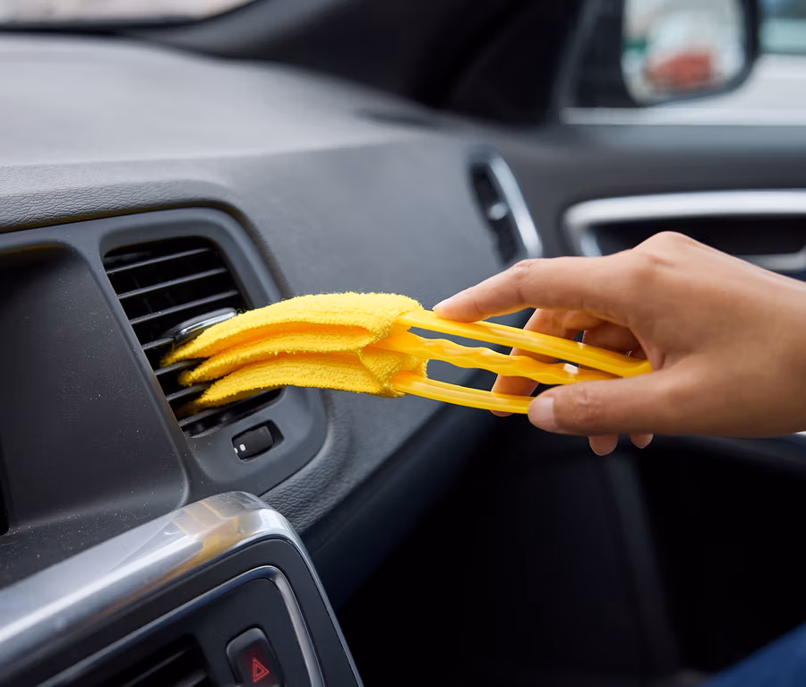 Eine Hand reinigt mit einem gelben Staubwedel eine Auto-Lüftungsöffnung.