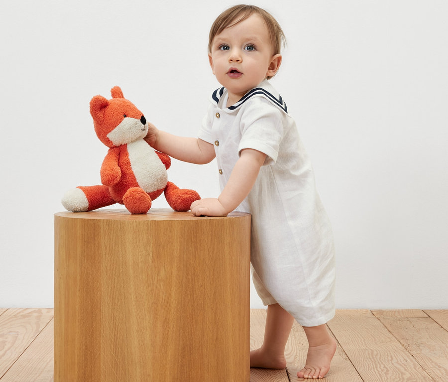 Baby in Matrosen-Strampler lehnt an einem Holztisch und hält einen Plüschfuchs.
