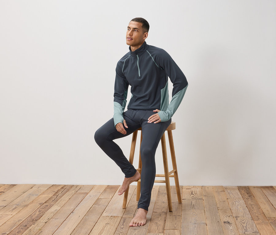 Ein Mann sitzt auf einem Holzhocker und trägt dunkelgraue Thermounterwäsche.