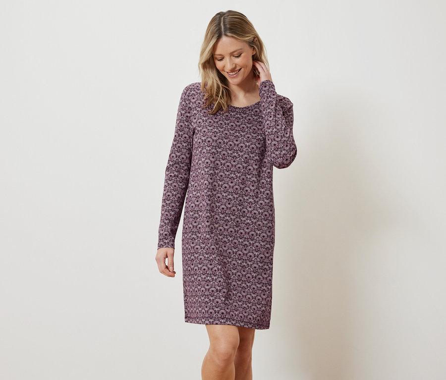 Eine blonde Frau lächelt und posiert in einem langärmligen Kleid mit Blumenmuster.