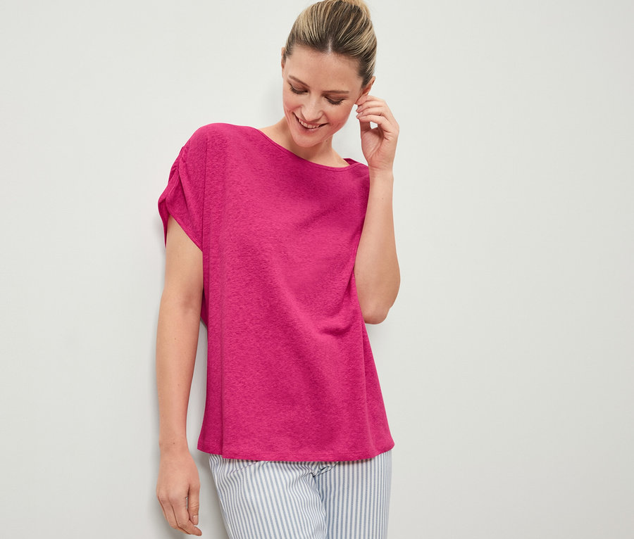Frau lächelt und berührt ihr Ohr, sie trägt ein rosafarbenes Shirt aus Leinenmix und eine gestreifte Hose.