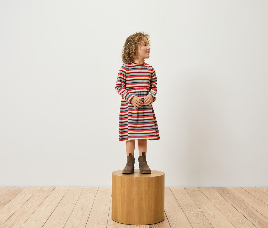 Mädchen mit lockigem Haar, bekleidet mit einem gestreiften Kleid und braunen Stiefeln, steht auf einem Holzhocker.