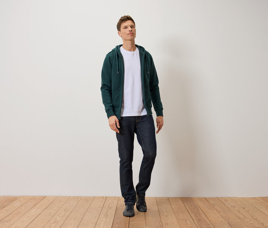 Mann steht auf Holzboden in dunkelblauen Jeans, weißem T-Shirt und grüner Reißverschlussjacke.