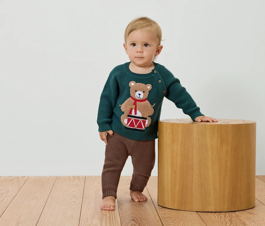 Ein Kind steht neben einem Holzhocker und trägt einen grünen Pullover mit einem Bären, der Trommel spielt, und braune Hosen.