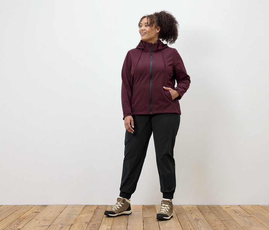 Eine Frau in einer burgunderroten Reißverschlussjacke, schwarzen Hosen und braunen Schuhen steht auf einem Holzboden.
