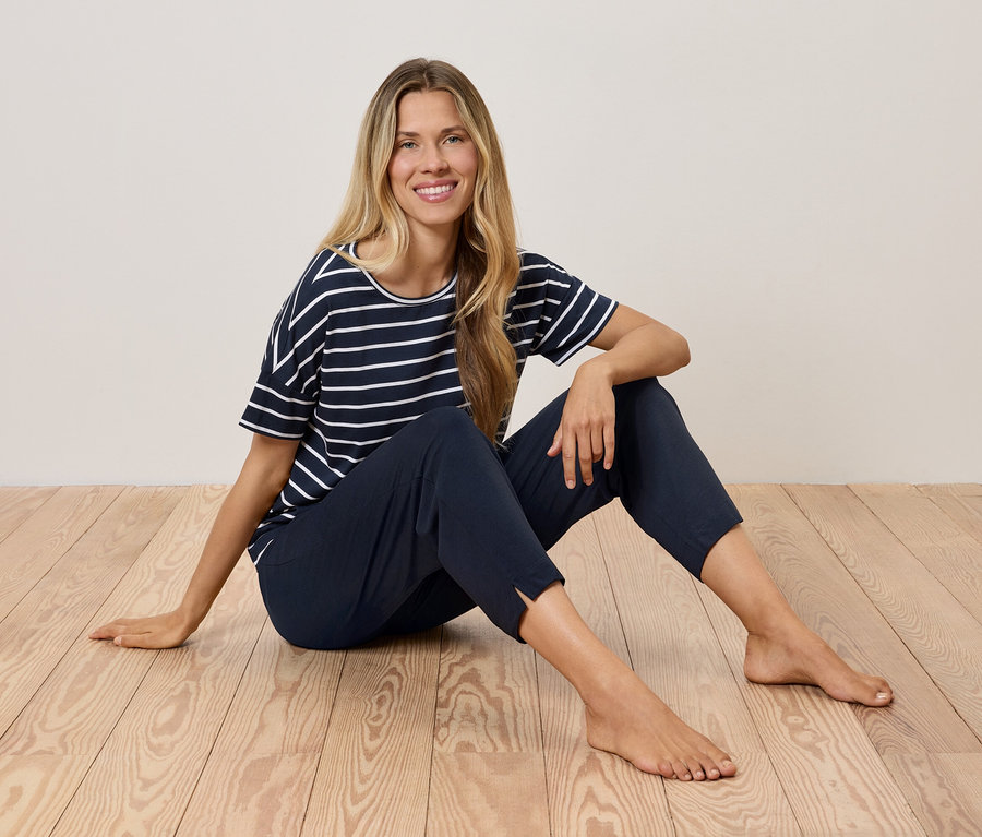 Blonde Frau sitzt auf einem Holzboden und trägt ein marineblau gestreiftes T-Shirt und dunkelblaue Hosen.