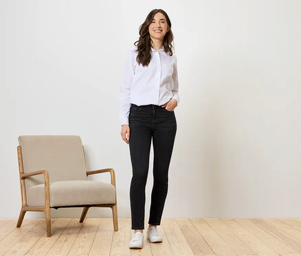 Frau steht in schwarzen Jeans, weißem Hemd und weißen Schuhen neben beigem Sessel.