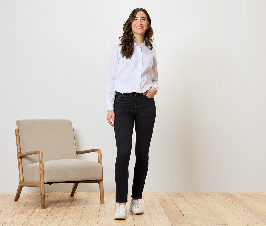 Frau steht in schwarzen Jeans, weißem Hemd und weißen Schuhen neben beigem Sessel.