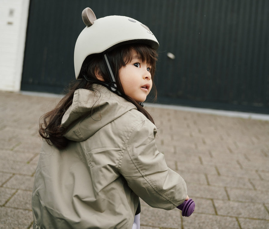 Mädchen mit braunen Haaren, beiger Jacke und blauen Jeans fährt auf einem lila Fahrrad und trägt einen HUDORA Kinderhelm, Beary.