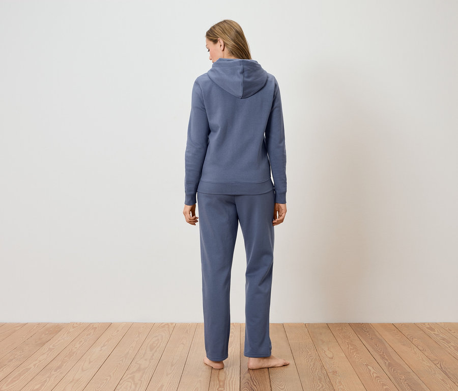 Rückansicht einer Frau, die einen blauen Hoodie und eine Hose vor einer weißen Wand trägt.
