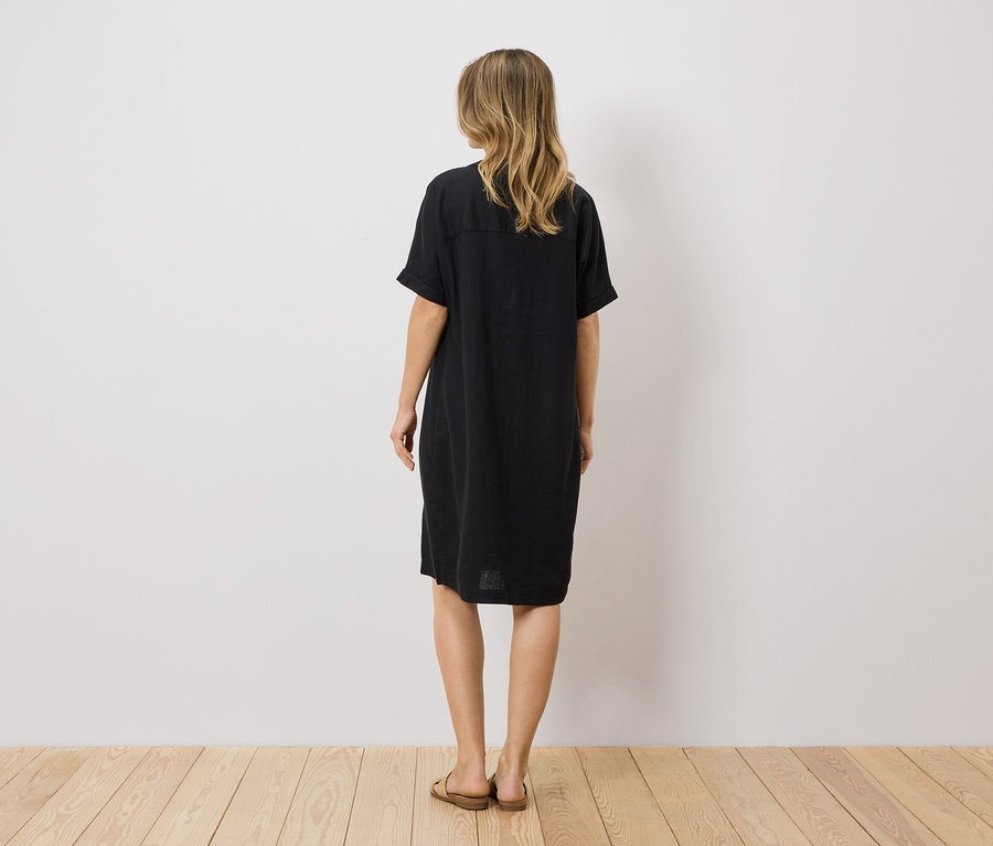 Rückansicht einer Frau mit blonden, gewellten Haaren, die ein schwarzes, knielanges T-Shirt-Kleid und braune Sandalen trägt.