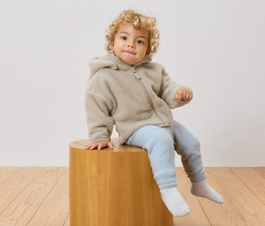 Ein Kleinkind mit lockigem Haar sitzt auf einem Holzhocker, trägt eine Fleecejacke mit Kapuze, hellblaue Hosen und weiße Socken.