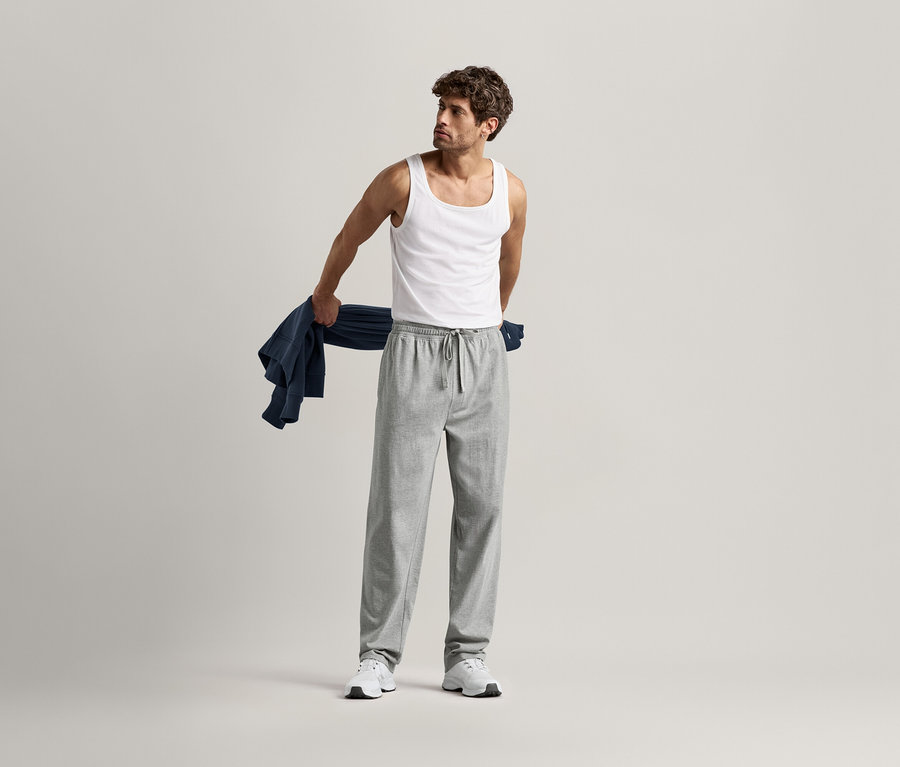 Mann im Unterhemd und grauer Relaxhose, hält ein blaues Hemd über dem Arm.