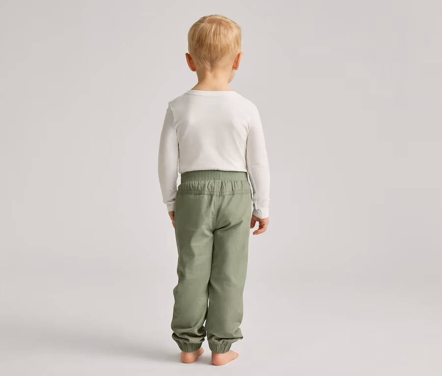 Ein Kind mit blonden Haaren steht mit dem Rücken zum Betrachter und trägt ein weißes Langarmshirt und grüne Hosen.