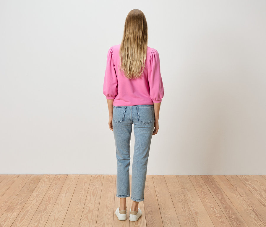 Rückansicht einer Frau mit blonden Haaren, die einen pinkfarbenen Pullover und hellblaue Jeans trägt.