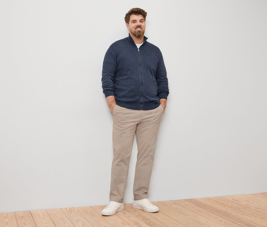 Ein Mann steht mit den Händen in den Hosentaschen, trägt eine marineblaue Strickjacke mit Reißverschluss, eine helle Hose und weiße Turnschuhe.