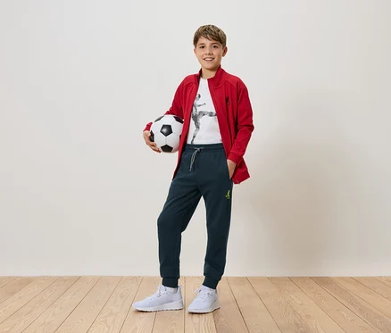 Ein Junge hält einen Fußball in den Händen. Er trägt eine rote Jacke, eine Hose und weiße Turnschuhe.