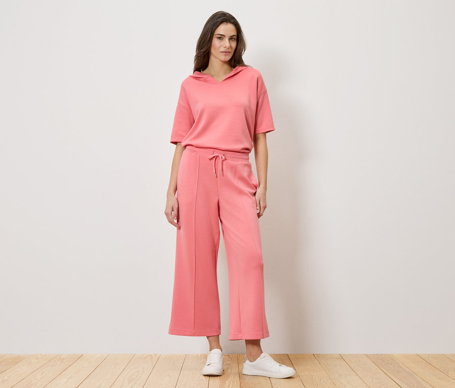 Frau steht vor einer Wand und trägt ein korallfarbenes, zweiteiliges Outfit, bestehend aus einem kurzärmeligen Hoodie und einer Culotte-Hose, zusammen mit weißen Turnschuhen.