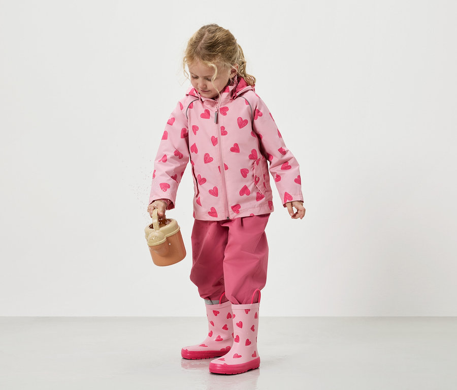 Ein kleines Mädchen schüttet Sand aus einem Spielzeugeimer, bekleidet mit einer rosa Jacke mit Herzmuster, einer passenden Hose und Gummistiefeln.