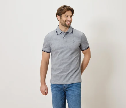 Mann mit grauem Poloshirt und Jeans, stehend vor einem neutralen Hintergrund.