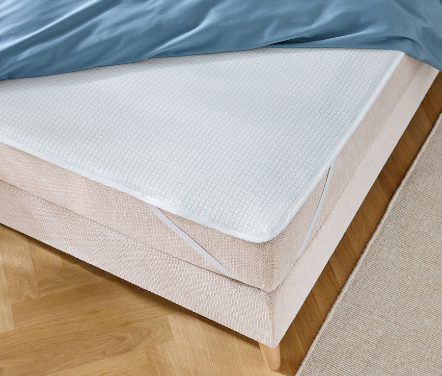 Nahaufnahme eines Bettes mit blauer Bettwäsche und einer weißen Matratzenauflage mit elastischen Bändern.